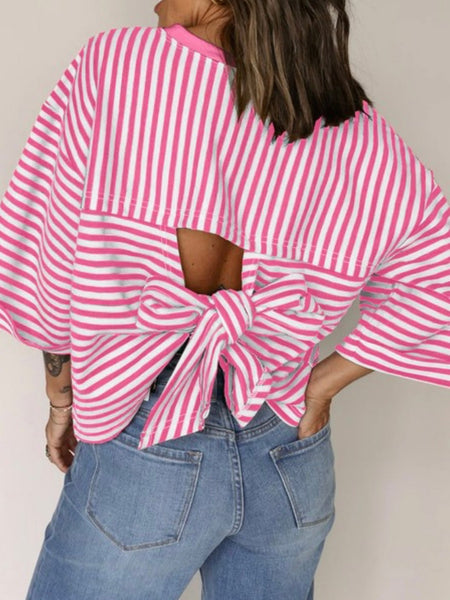 Woman wearing a pink and white striped top with open back and oversized bow tie detail, paired with denim jeans.