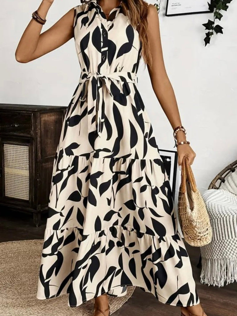 Sleeveless black and cream printed maxi dress with belted waist and tiered skirt, styled with a straw tote bag.